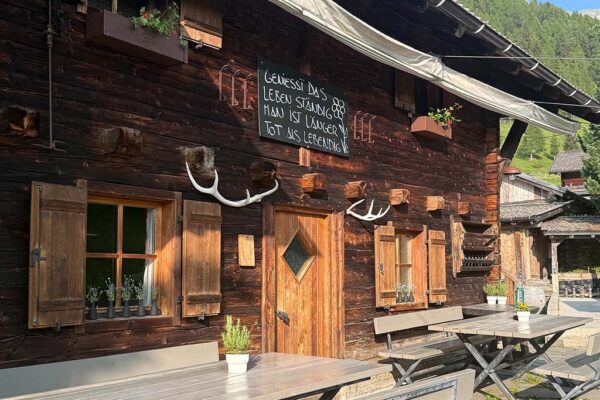 Foto der Frontfassade der Alm-Hütte mit möblierter Terasse und Bergen im Hintergrund.