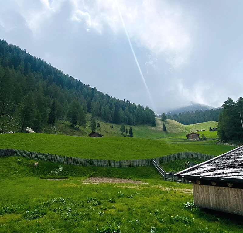 Grüne Almwiese mit Holzhütten und Zaun, umgeben von bewaldeten Bergen, während Sonnenstrahlen durch die Wolken auf die Landschaft fallen.