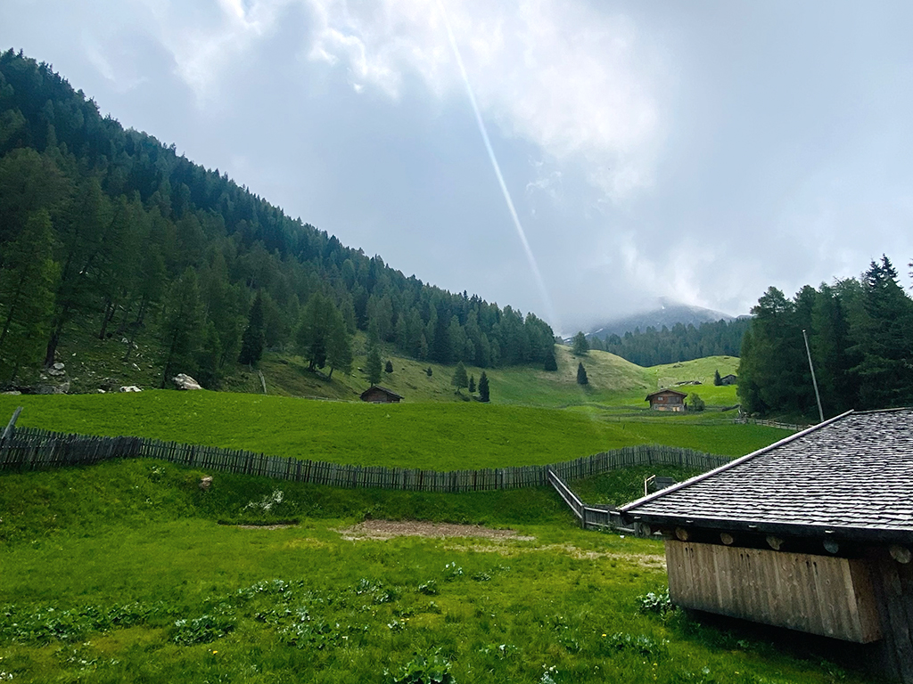 Grüne Almwiese mit Holzhütten und Zaun, umgeben von bewaldeten Bergen, während Sonnenstrahlen durch die Wolken auf die Landschaft fallen.